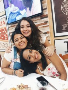Tres mulheres em uma pizzaria, a cima uma tv de tela plana, abaixo uma mulher de cabelos pretos de pele clara sorrindo, logo a baixo uma menina de cabelo preso, sorrindo de macacão jeans e abaixo dela uma menina sorrindo de cabelos soltos, de vestido branco com corações rosa.