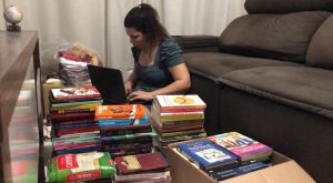 Da esquerda para direita. mesa marrom de mogno, logo a direita vemos pilhas de livros e uma mulher com uma blusa cinza utilizando um notebook preto, atrás um sofá retrátil marrom